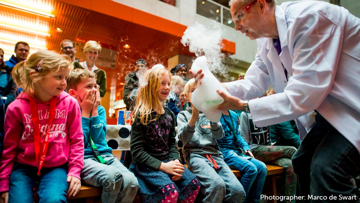 Mad scientist showing a beaker with smoke and bubble poring out to children while parents look on