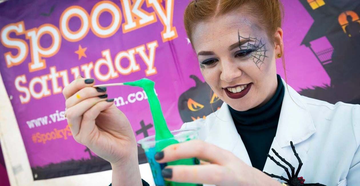 A mad scientist with a spider web drawn on her face playing with green slime in a cup